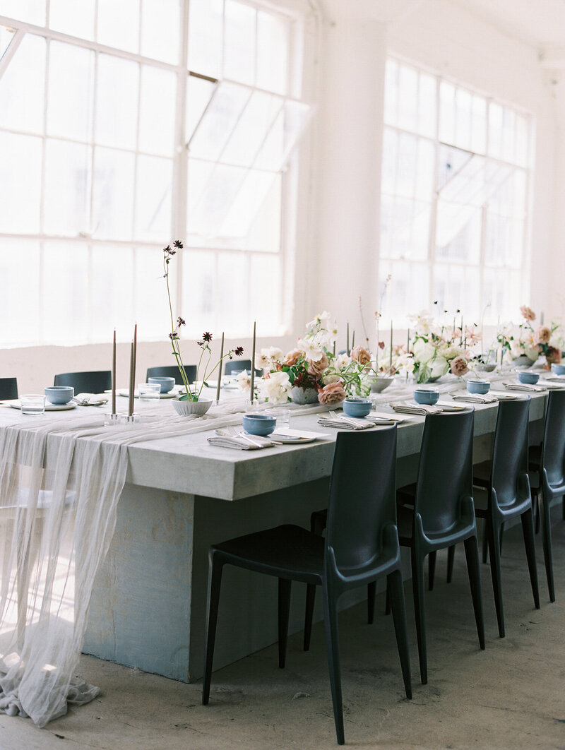 A wedding place-setting, complete with three stacked white plates, each smaller than the last. With Golden yellow cloth napkins, silver cutlery, various sized drinking glasses, and a short pink and green floral arrangement in a round soft-stone vase, with a short and skinny stem.