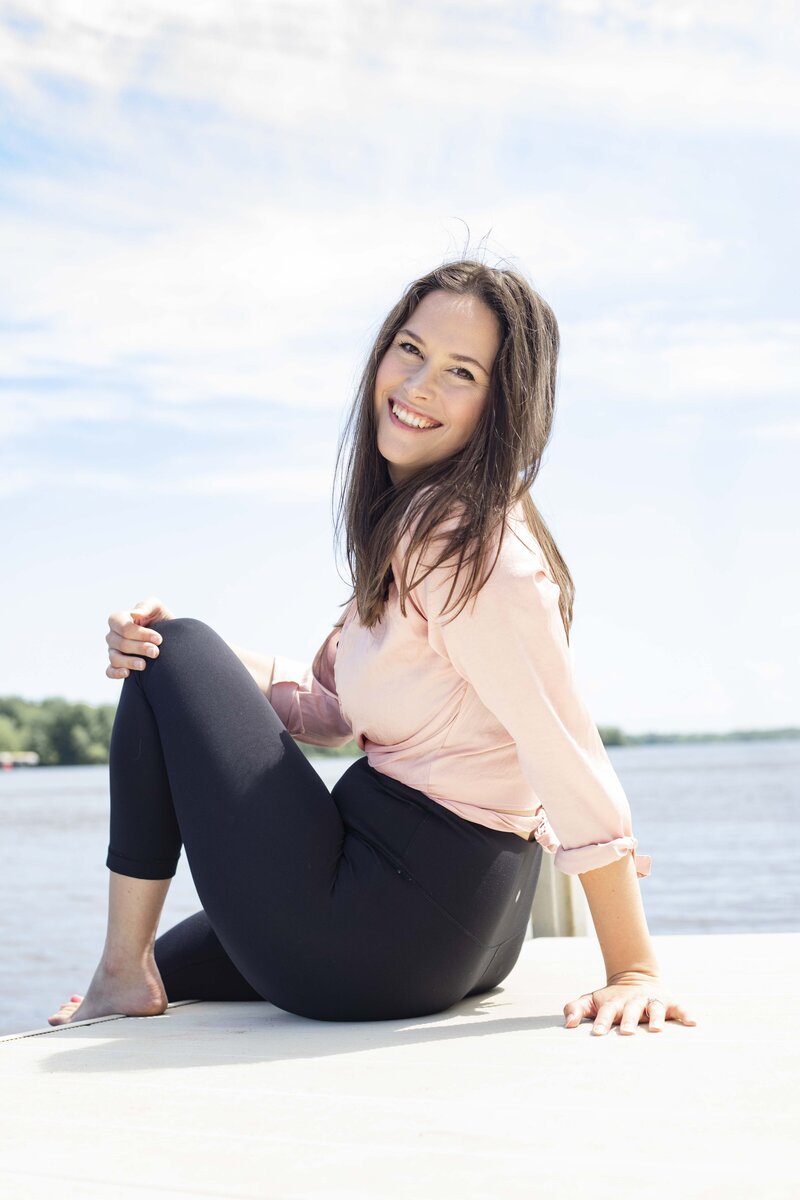 Cassie Schmidt of Creative Business Coach sitting on a lake pier in black leggings and a pink top.