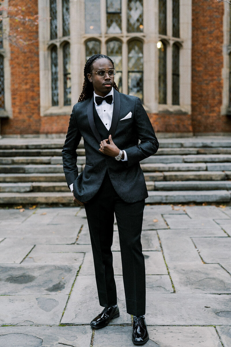 Man wearing a black tuxedo standing confidently on stone steps outdoors