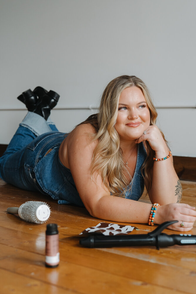 A young lady is laying on the floor in a studio. There are hair coloring items and a curling iron in the floor in front of her. She is wearing a denim outfit and smiling off to the side. Branding photos for hair stylist in Elizabethton, TN. 