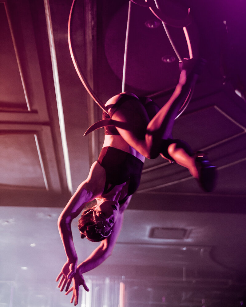 An aerial hoop performer hangs upside down at a party, lit in purple.