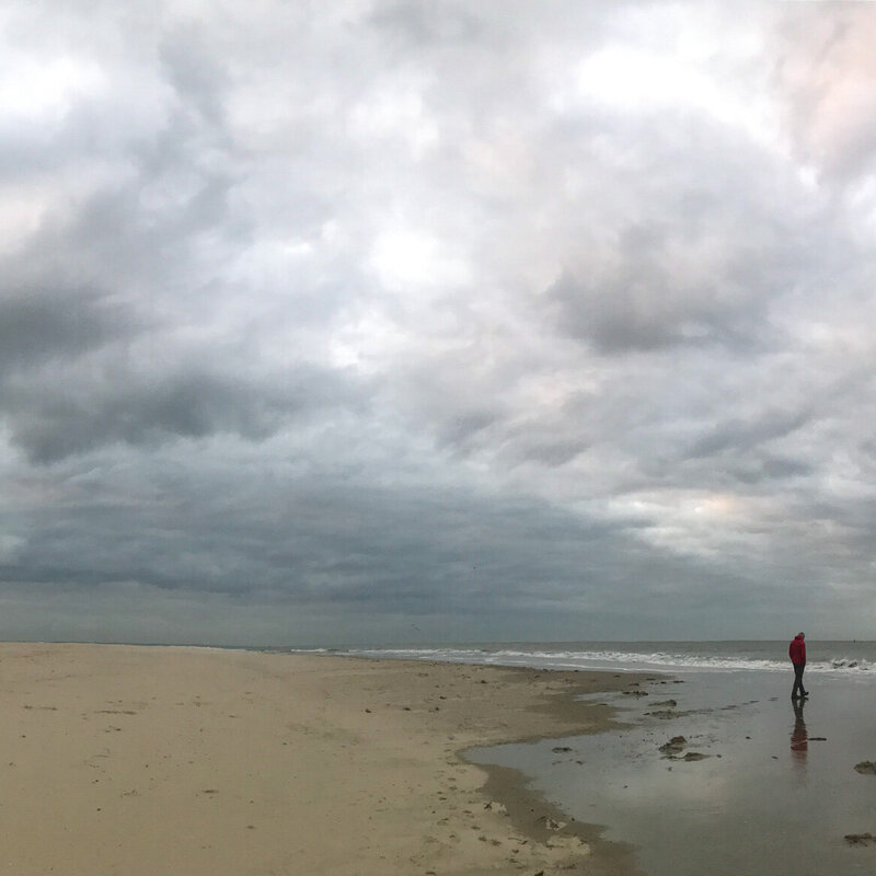 A man deep in thought wearing a red coat walking towards the waves on a stormy beach