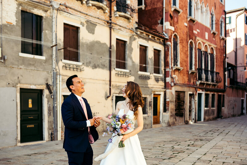 The bride and groom share a lighthearted moment, laughing as they take photos amidst the charming and winding alleys of Venice, creating joyful memories on their wedding day