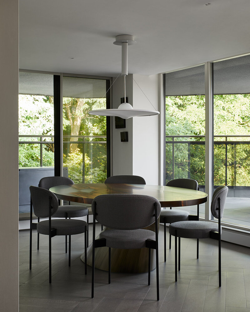 A spacious dining room featuring a large, round wood dining table surrounded by soft grey chairs, a diffused globe pendant light, and tall windows