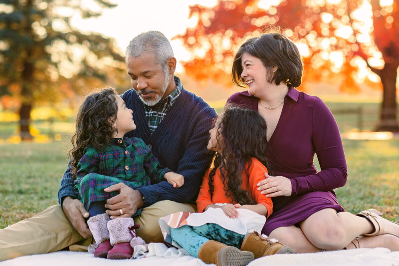 A family of four sitting on a blanket and smiling at each other during their fall photo session in Burke, Virginia.