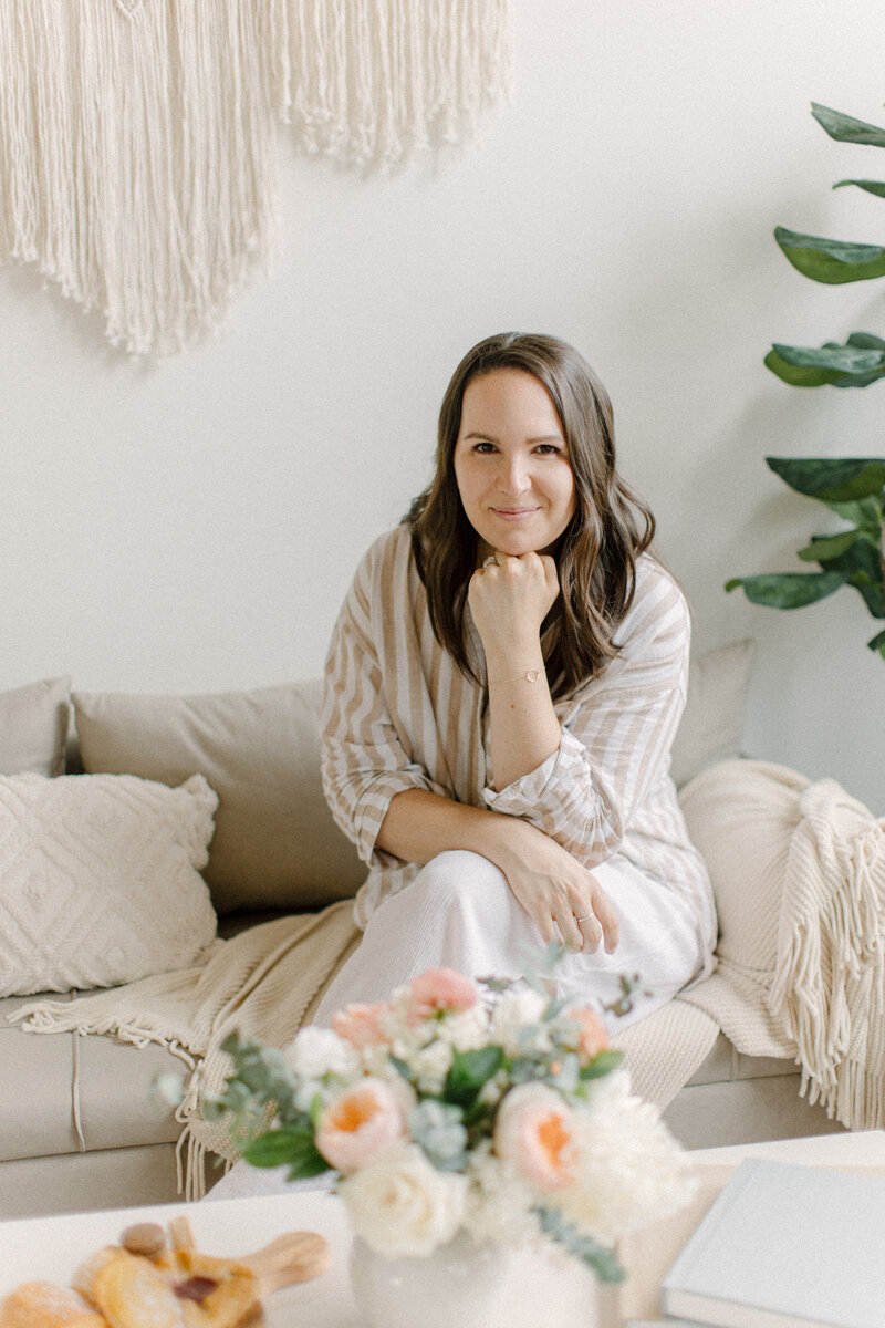 Boston motherhood photographer, Corinne Isabelle is wearing a light beige and white striped shirt, white pants and is sitting on a couch