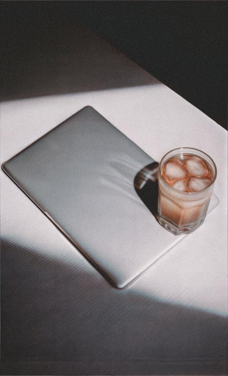 Minimalist flatlay of a closed laptop and iced coffee casting soft shadows in natural sunlight — peaceful and modern workspace for a soulful entrepreneur