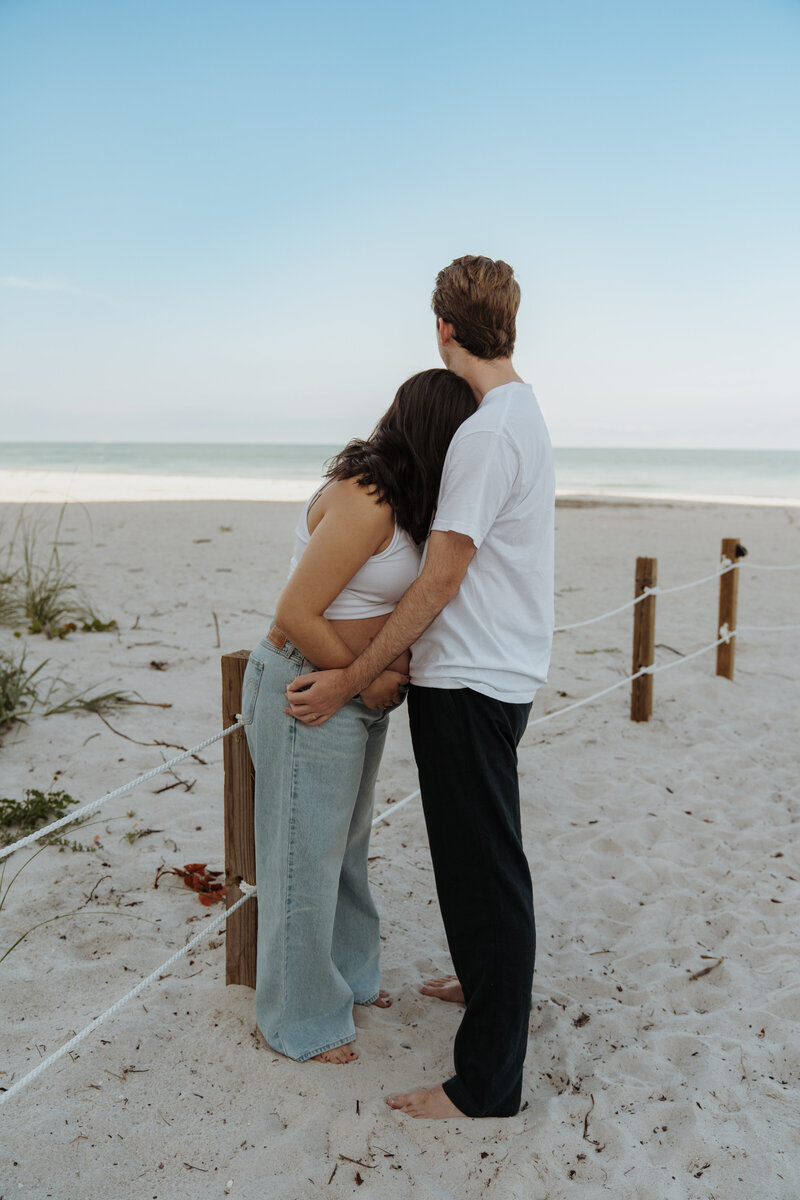 Couple standing barefoot on the sandy beach with the woman leaning her head on the man’s shoulder, both wearing white tops and looking out over the ocean horizon.