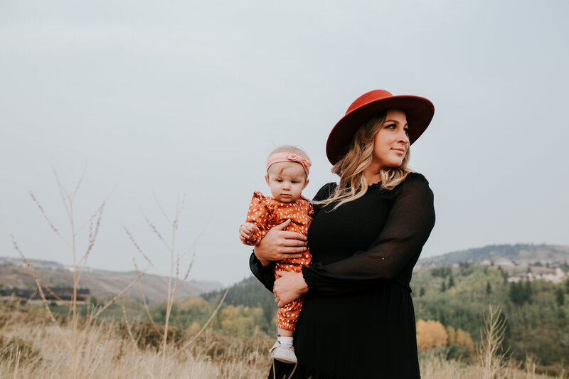 A young mom in a black dress and red hat holds her baby daughter on her hip while looking off to the side with the Alberta foothills in the backgroun. her daughter in wearing a red polkadot romper with a pink bow headband in her hair