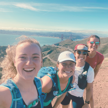 Four women in running gear smile at the camera on a brown dirt trail. In the background you can see the San Francisco Bay, and the tops of the Golden Gate Bridge, with San Francisco in the distance. The sky is blue with wispy white clouds.