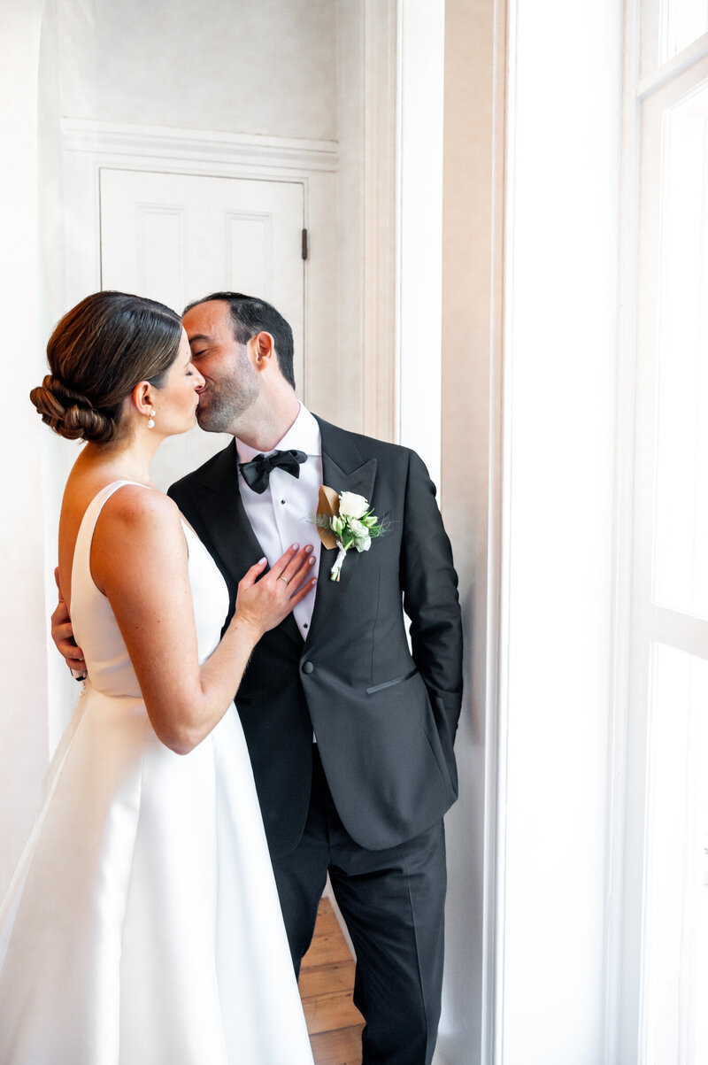 A husband and wife kissing on their wedding day inside of a white house.