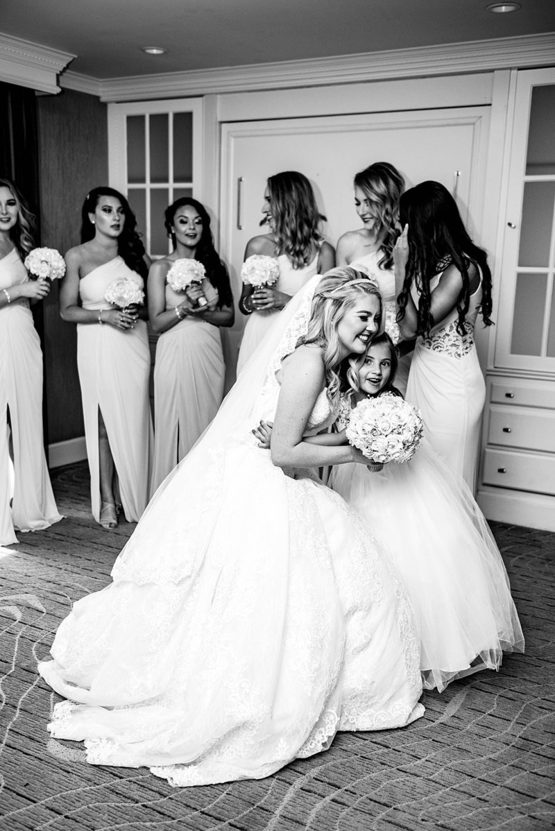 Black and white photo of flower girl hugging the bride after she gets into her wedding dress, bridesmaids standing behind them smiling