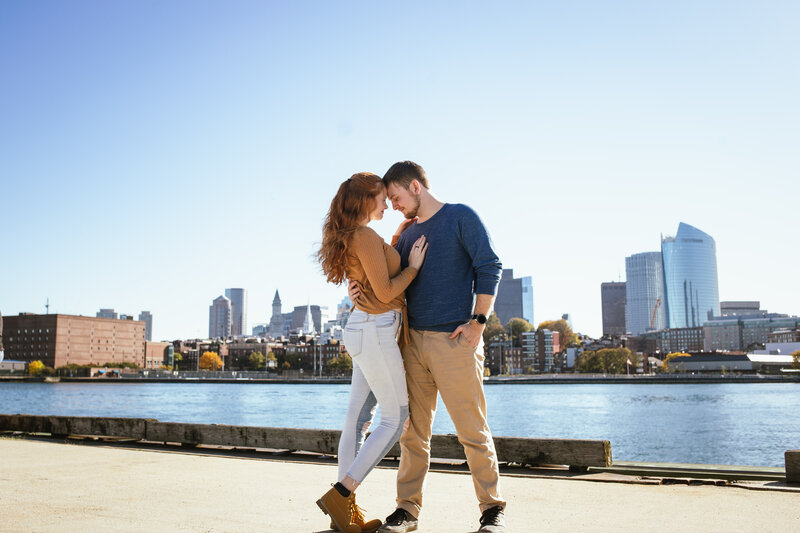 A couple rest their foreheads together and cuddle with the Boston bay and skyline behind them.