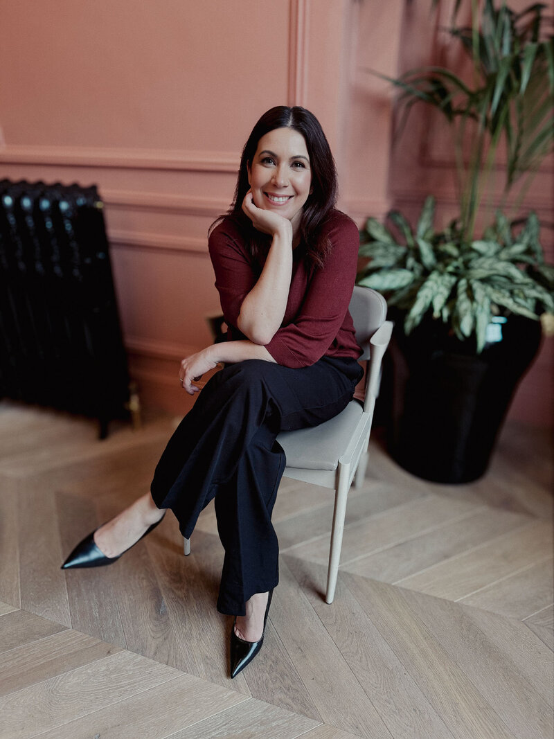 Roberta Rechena from ByRoberta Design sits on a chair with legs crossed and chin in one hand as she smiles at the camera. Roberta wears a wine coloured top and black trousers.