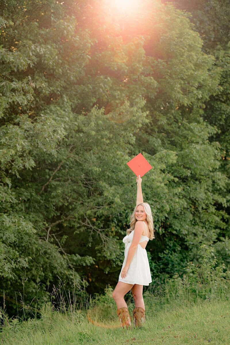 a blond charlotte area grad holds her orange graduation cap in the air while standing in a grassy field