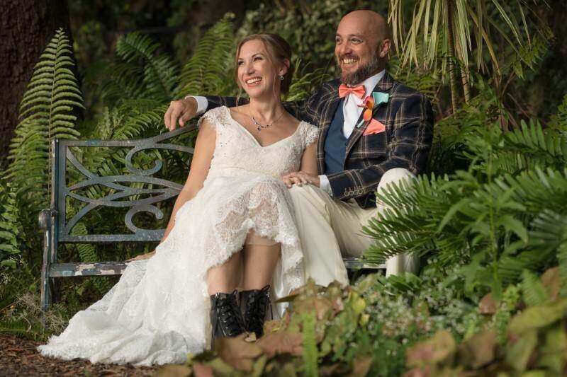 A joyful couple in wedding attire sits on a garden bench surrounded by lush greenery and smiling brightly