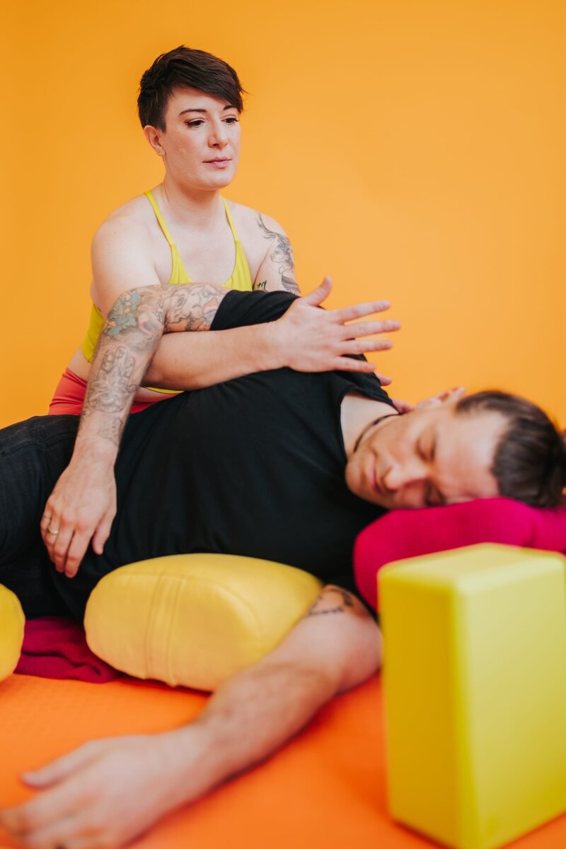 Massage therapist holds man in a stretch as he lays on colorful bolsters with a yellow-orange backdrop,