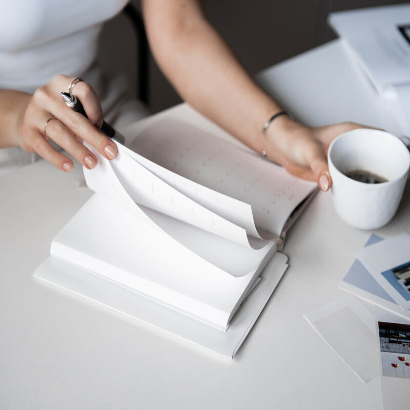 woman's hand flipping through notebook while sitting at desk and holding a white  cup with coffee