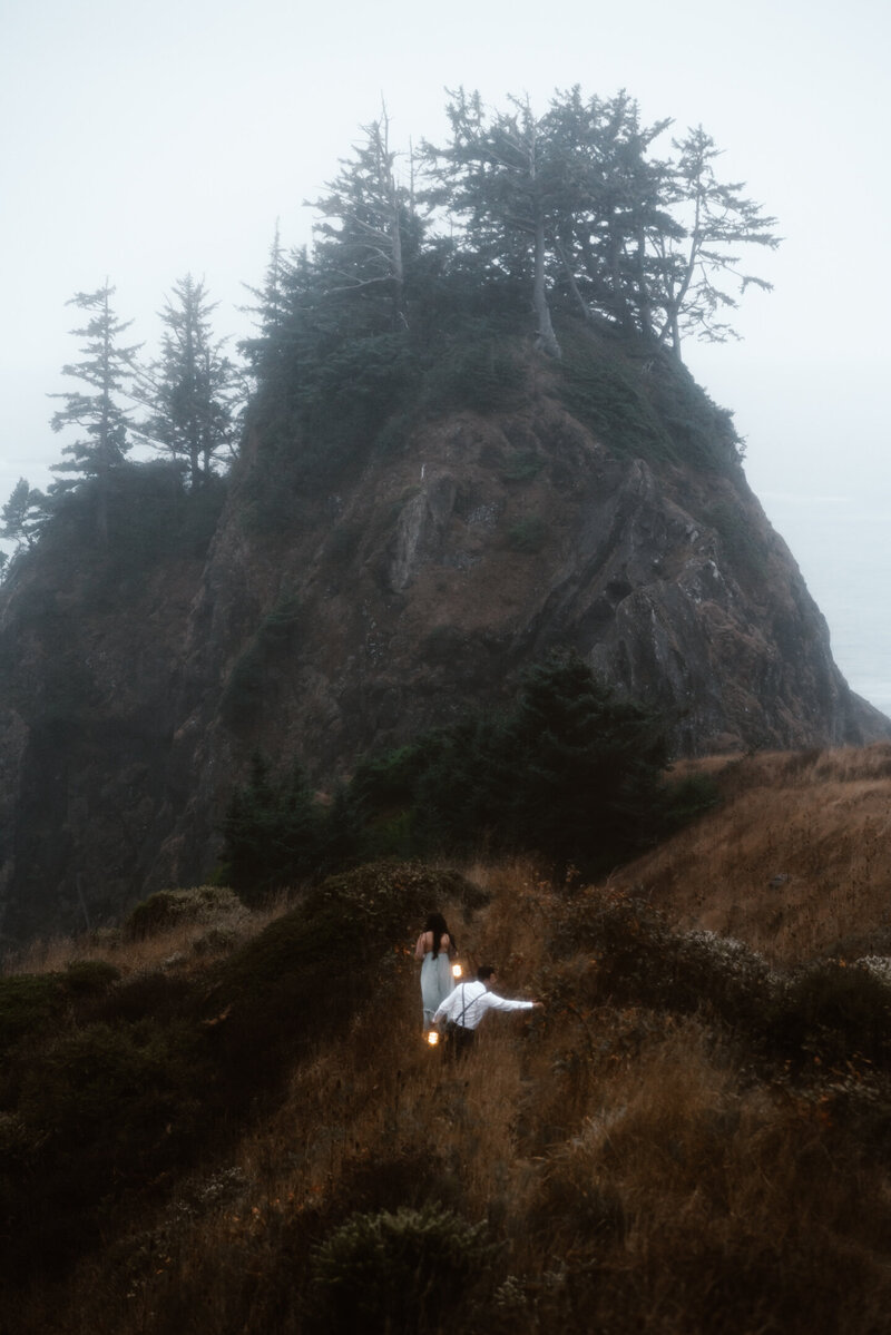 During a moody Oregon coast elopement, a bride and groom explore a cliffside location. it is barely morning and a heavy fog surrounds them. The use lanterns to light their path.