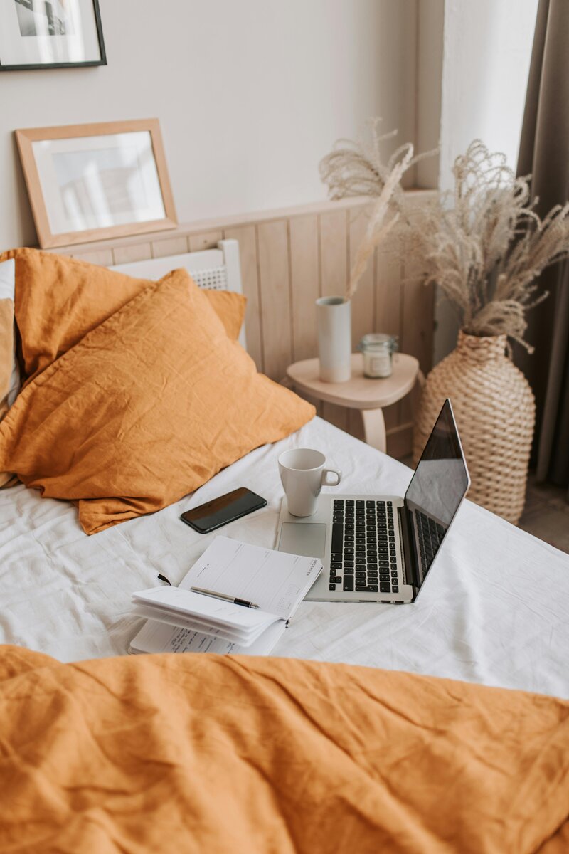 Image of a bed with a laptop, coffee, agenda, and phone in a work from home set up