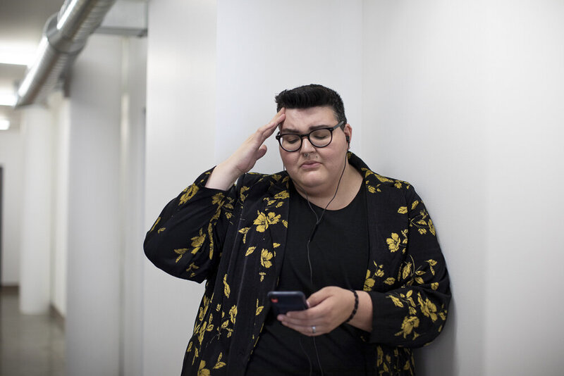 This image shows a feminine-presenting person with short, cropped hair and light skin, wearing a black and yellow floral blazer. They stare down at their phone in their left hand, with their right hand pressed to their forehead, a concern expression on their face.
