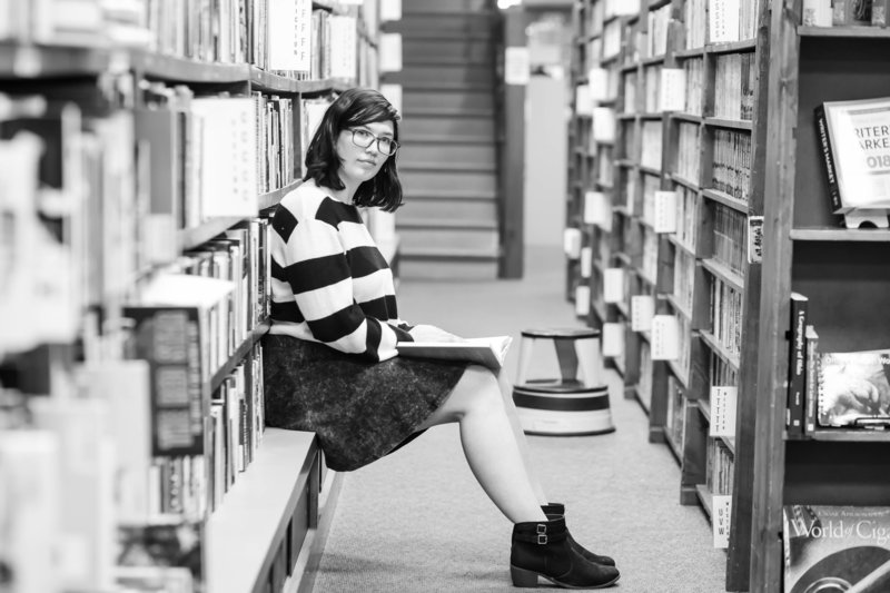 waynedale high school senior girl sitting on a book shelf ledge in books in stock wooster ohio, with books on her lap looking seriously towards camera, black and white photo, she was wearing black and white stripped shirt with black skirt, photographed by Jamie Lynette Photography canton ohio senior photographer