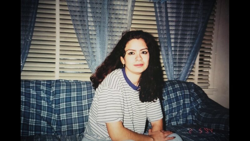 A woman with dark, wavy hair sits on a plaid couch, wearing a striped t-shirt. Behind her are closed blinds and sheer blue curtains. The date '2-5-94' is displayed in the bottom corner, suggesting the photo was taken in 1994.