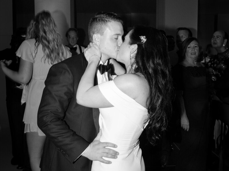 A couple in formal attire shares a kiss at a celebration, surrounded by guests. The woman wears an off-shoulder dress, and the man is in a suit. The background shows people in semi-formal outfits enjoying the event.