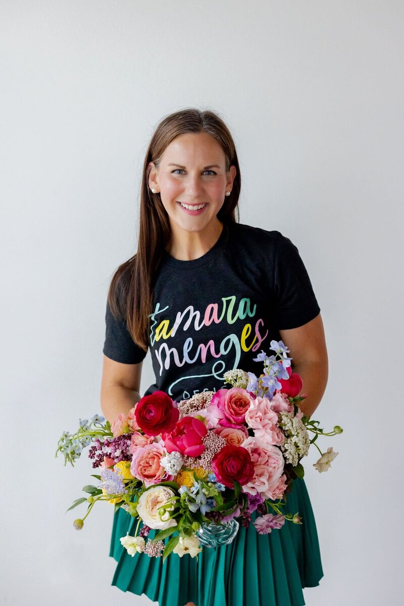 Tamara Menges holding a vibrant garden-style bridal bouquet with colorful blooms, smiling in studio with floral arrangements.