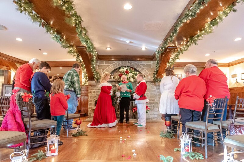 A indoor Christmas-themed wedding ceremony at Chetola Resort and Spa in Blowing Rock, NC with a bride in a red holiday gown and groom in a Santa-inspired suit, standing at the altar surrounded by family members in festive clothing under holiday garlands and lights.