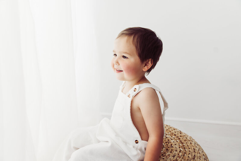 A young child with short dark hair in a white outfit sits on a wicker stool, looking toward a sunlit window with a joyful expression. The background is bright and minimalistic, creating a serene atmosphere.