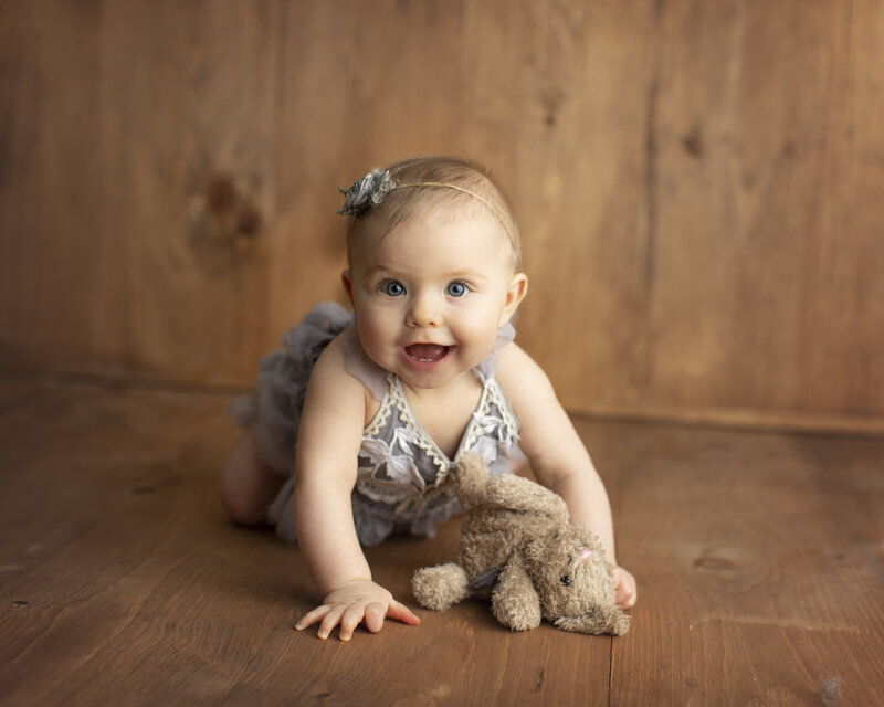 Baby girl smiles at camera while crawling on wooden floor for her milestone session.