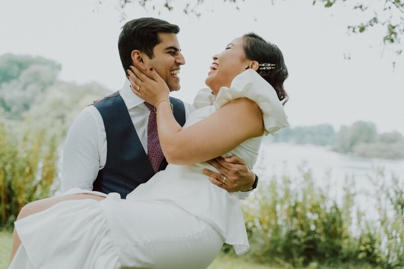  A romantic portrait of a couple standing on a wooden deck by a lake. The man, in a dark blue suit with a purple tie, lovingly kisses the forehead of the woman, who is wearing a white dress with frilly sleeves. In the background, a windmill stands on the far side of the water under a cloudy sky.