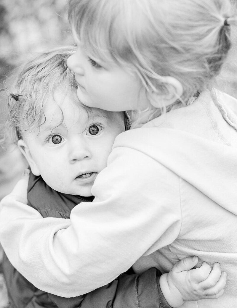 Little brother and sister hugging each other, with the brother’s eyes wide open, for light and airy photography.