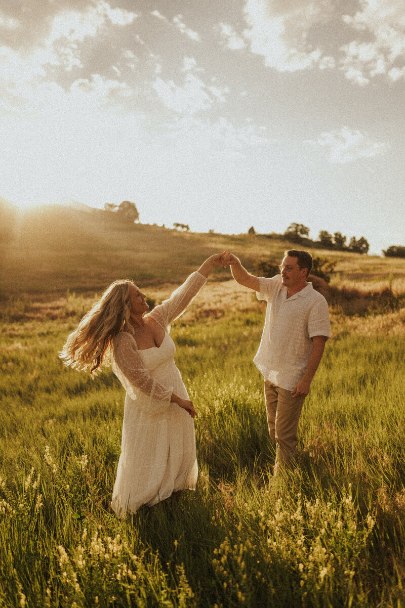 A couple dances in a golden field at sunset, with one person twirling the other while smiling, capturing a moment of joy.