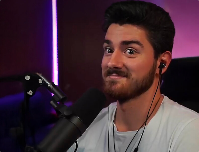 Man sitting in a purple-lit room with short brown hair and full beard and mustache. He is smiling slightly and wearing a white shirt. A microphone is propped next to his mouth.