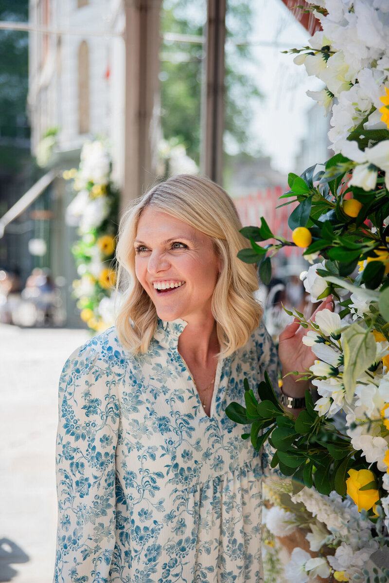 Emily Collins smiling while standing next to a floral display with white and yellow flowers, wearing a patterned blue dress
