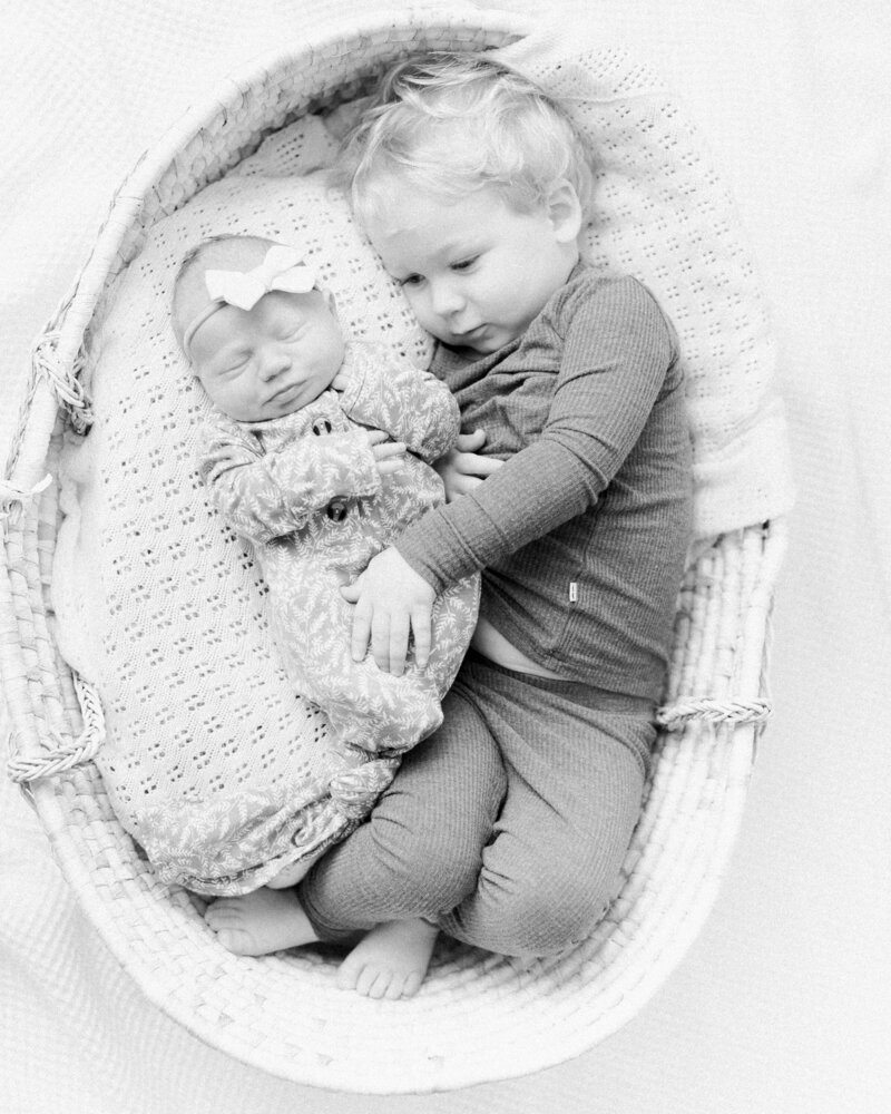 New big brother snuggles next to his newborn baby sister in a moses basket, and softly soothes her, by Boston family photographer Fieldstone Studio.