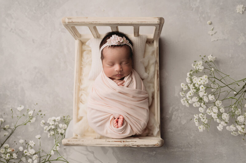 sleeping newborn baby girl swaddled in a pale pink swaddle with her toes peeking out, sleeping in a tiny white bed surrounded by baby's breath