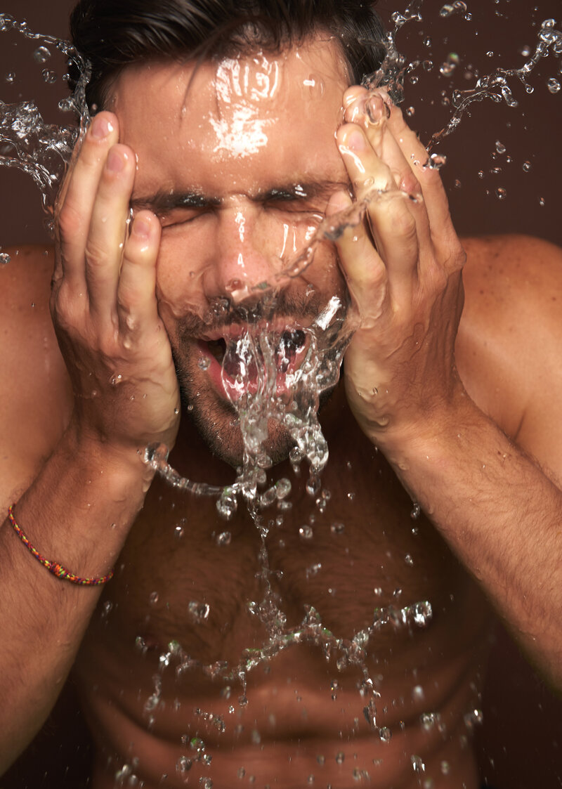 Attractive shirtless white man with smooth skin and beard washing face with a brown background.