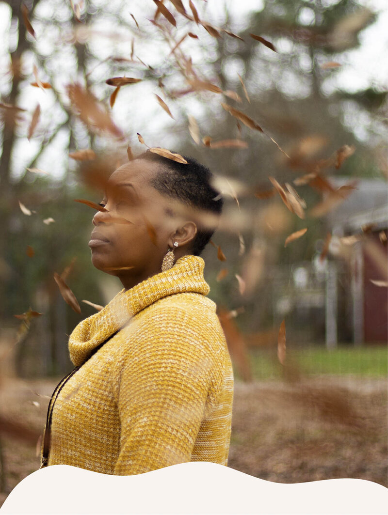 A burst of blowing fall leaves are visible in the foreground. In the background, a feminine presenting person of color looks past the viewer, into the distance, wearing a yellow sweater and textured jewelry. The photo is framed by a snow-like design.