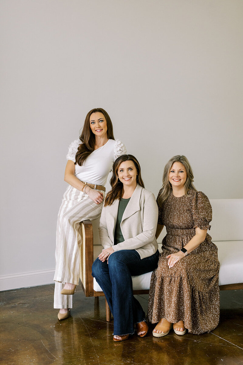 Three women are posing indoors against a plain light-colored wall. One stands on the left wearing a white top and striped pants. The other two are seated; the middle one in a beige jacket and jeans, and the right one in a brown dress.