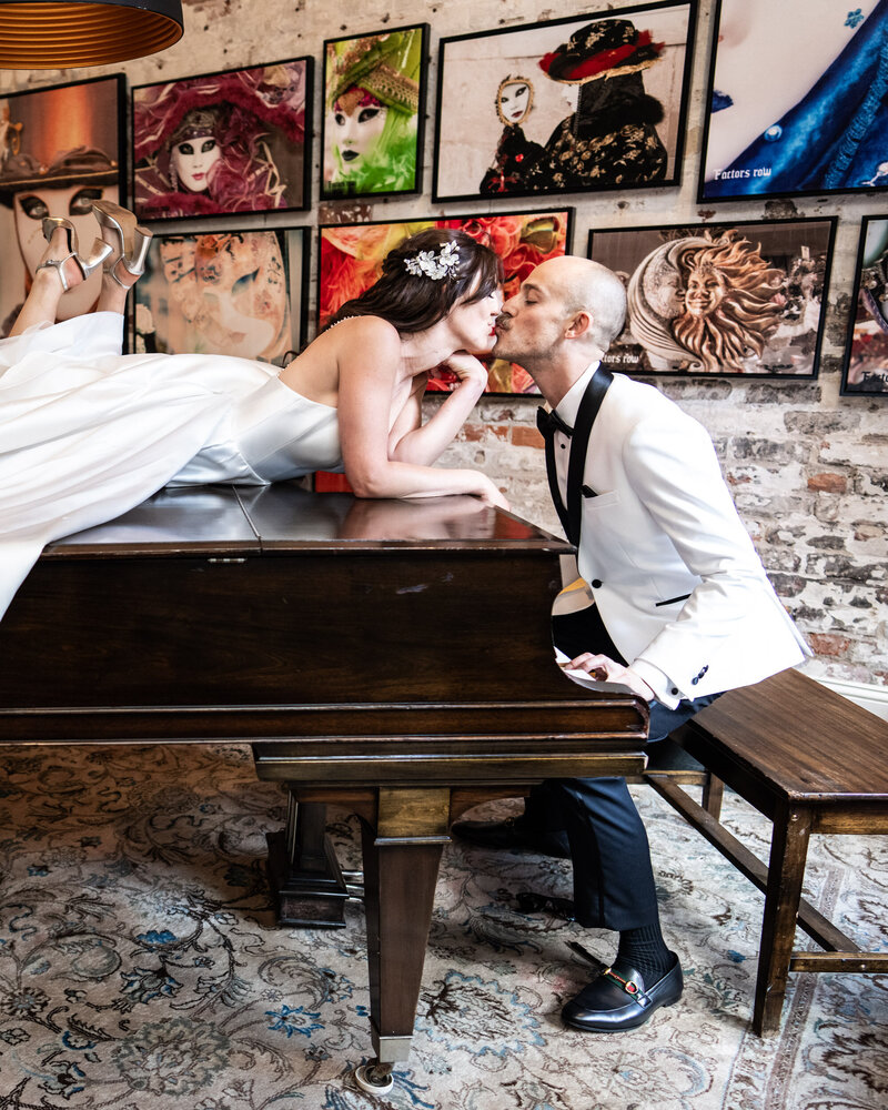Bride on top of piano kisses seated groom, with artwork in the background