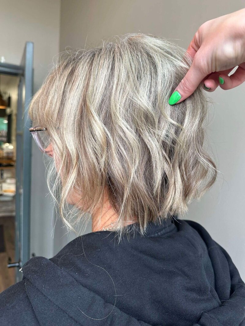 A woman with a textured, wavy blonde bob hairstyle, with a hand gently adjusting her hair, showcasing a modern cut created by 212 Salon, Spa, & Barbershop.