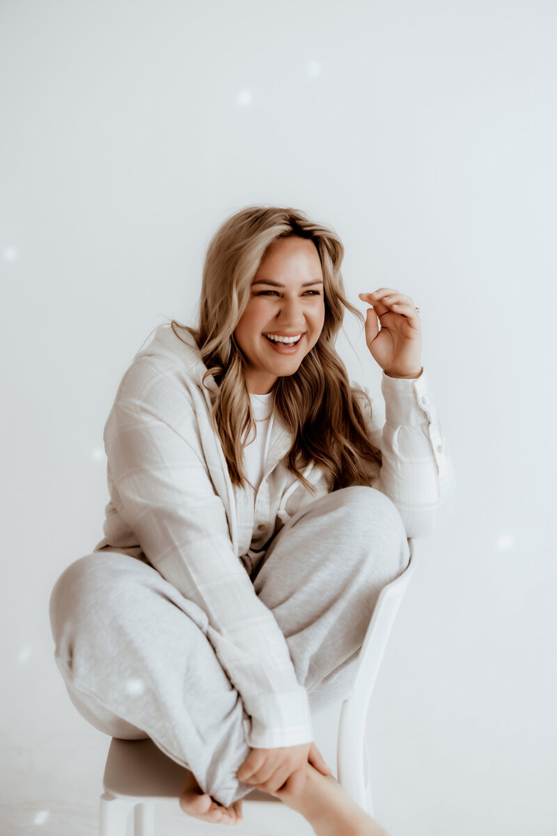 Photographer in Denver poses for a headshot in a white sweat suit while laughing against a white backdrop