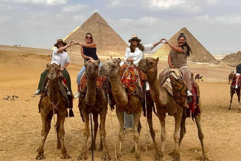 Terra from Adventures by Terra with group of 3 other women sitting on camels making heart signs with their hands with the pyramids of Giza in the background.