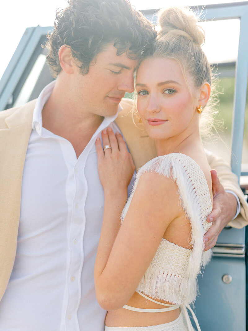 Engaged couple embracing while leaning on a blue Ford Bronco with the dunes of Nantucket behind them.