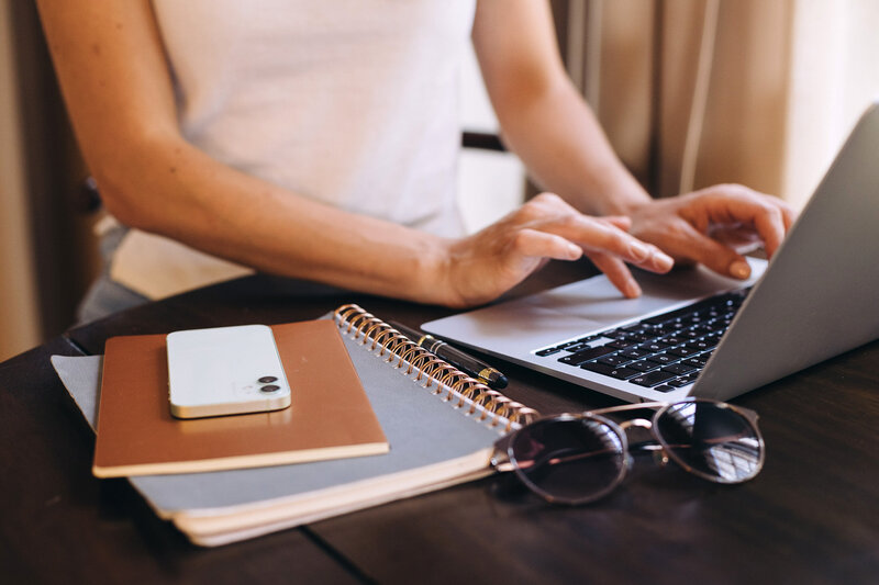 Close-up of a person typing on a laptop at a desk with a spiral notebook, pen, and sunglasses in the foreground. This image supports messaging about Fractional COO services that bring strategic clarity, operational structure, and long-term growth to busy architects and interior designers. 