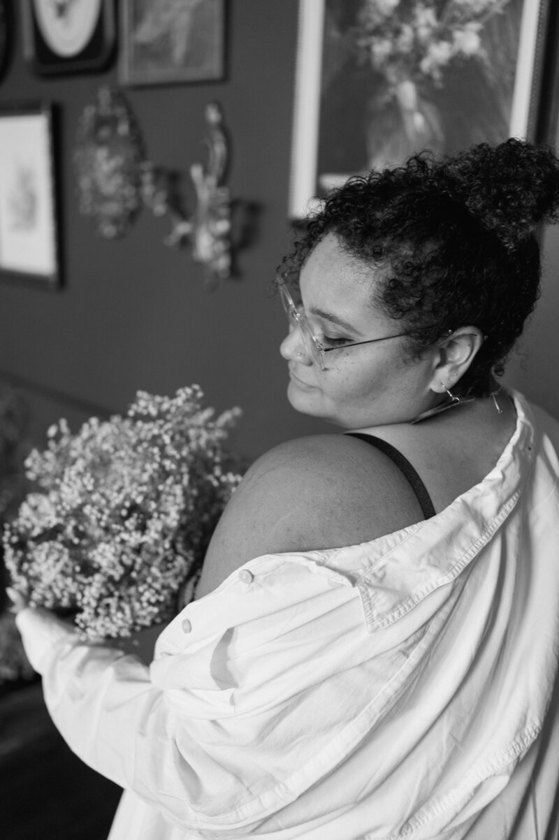 Ashley Vaughn looking over her shoulder, she is holding baby's breath with a lace black strap body suite, and a white over-sized shirt falling off her shoulder. She has curly hair, in a bun, clear glasses, nose peircings, and lots of ear piercings.