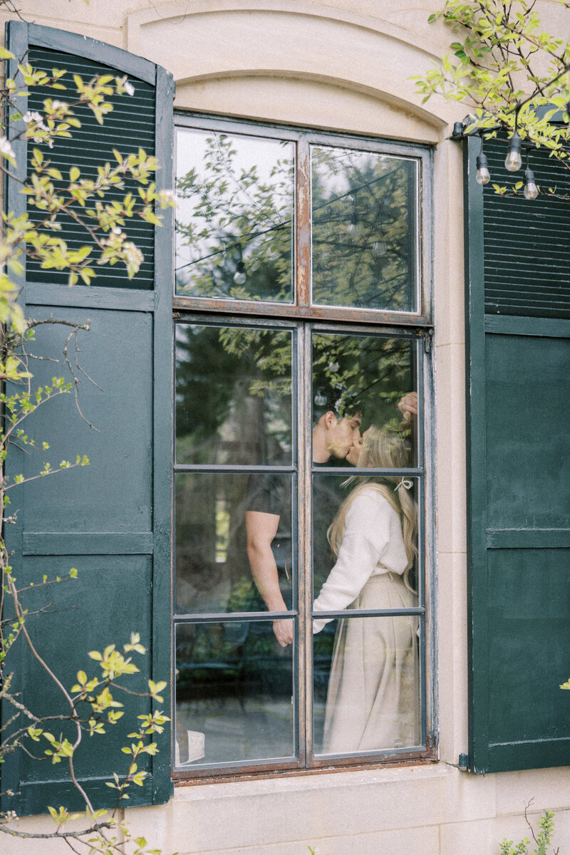 Couple standing in a window  sharing a kiss in a classic white engagment session dress